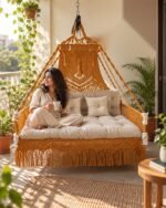 Woman relaxing on a mustard macrame sofa swing jhula with ivory cushions on a sunny balcony
